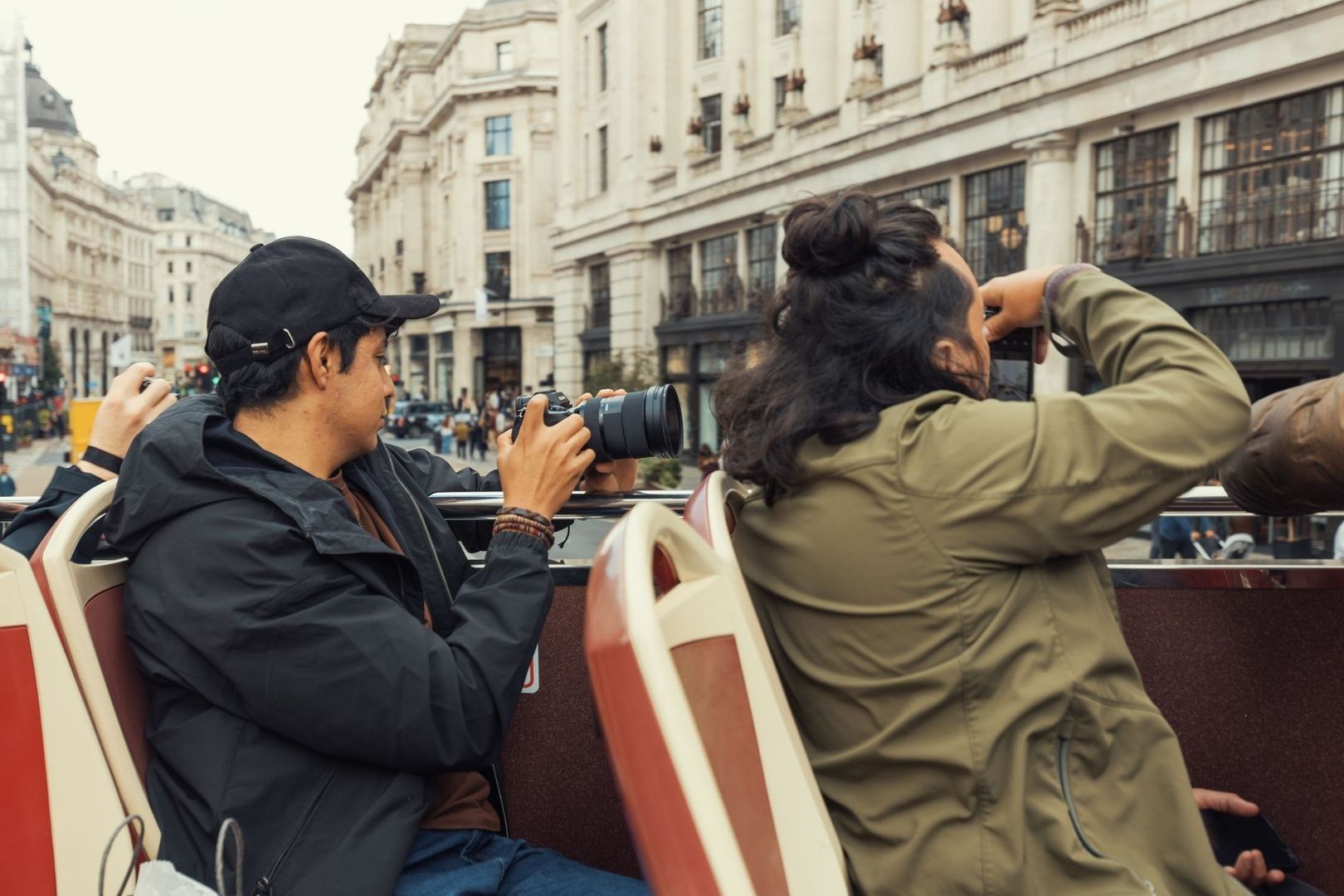Two people photograph city buildings from an open-top tour bus